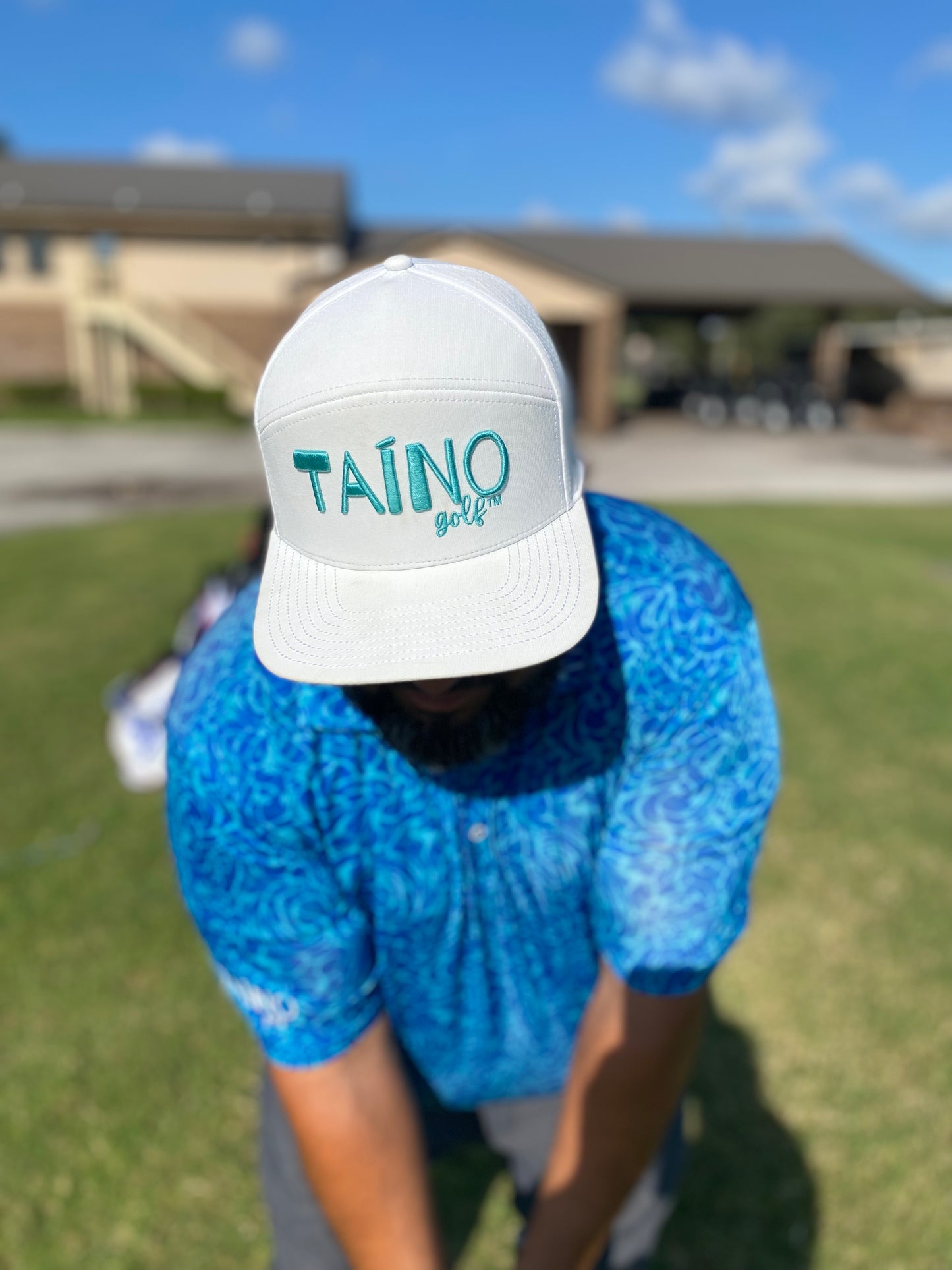 Golfer with a white Taino Golf hat with teal logo embroidered on the front, chipping at a golf course.